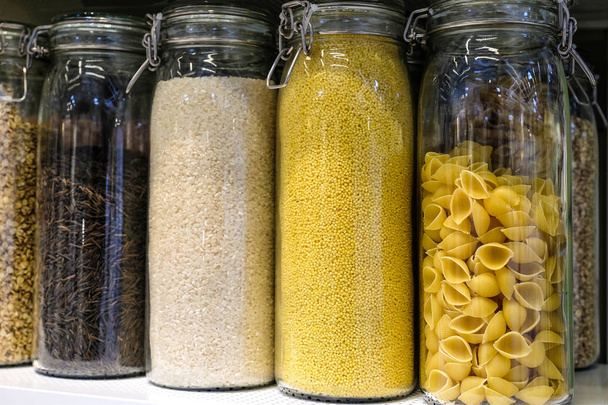 Various raw cereals, grains, beans and pasta for healthy cooking in glass jars on kitchen shelf, white background. Clean eating, vegetarian, vegan, balanced dieting food concept - Φωτογραφία, εικόνα