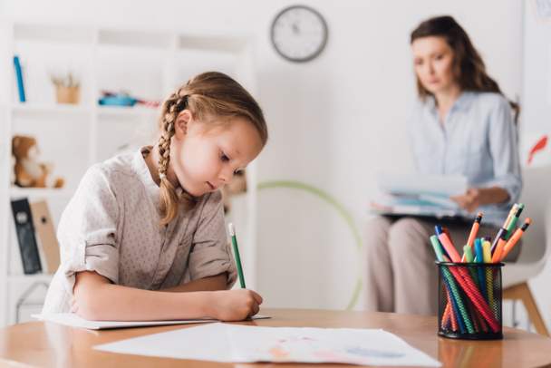 serious little child drawing while psychologist sitting blurred on background - Fotoğraf, Görsel