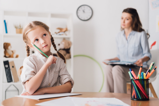 thoughtful little child drawing while psychologist sitting blurred on background - Photo, Image