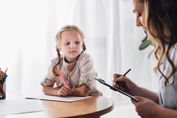 Psychologe schreibt in Klemmbrett, während er in der Nähe eines kleinen zeichnenden Kindes sitzt - Foto, Bild