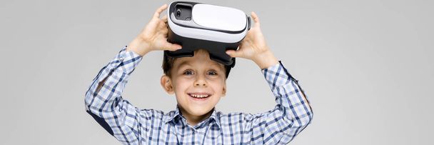 A charming boy with an inlaid shirt and light jeans stands on a gray background. The boy on his face glasses virtual reality - Photo, Image