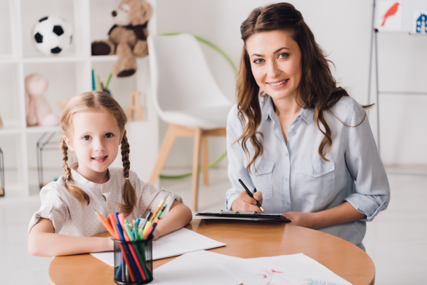 psicólogo sonriente con portapapeles sentado cerca de un niño pequeño mientras ella dibuja con lápices de color y mirando a la cámara
 - Foto, imagen