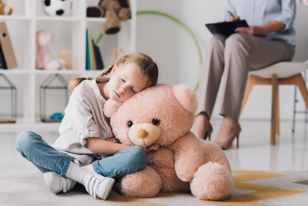 lonely little child with teddy bear sitting on floor with psychologist sitting on background - Photo, Image