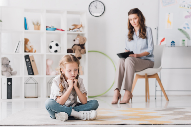 depressed little child sitting on floor with psychologist sitting on background - Foto, imagen