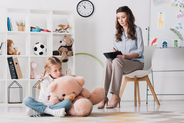 depressed little child with teddy bear sitting on floor with psychologist sitting on background - Photo, Image