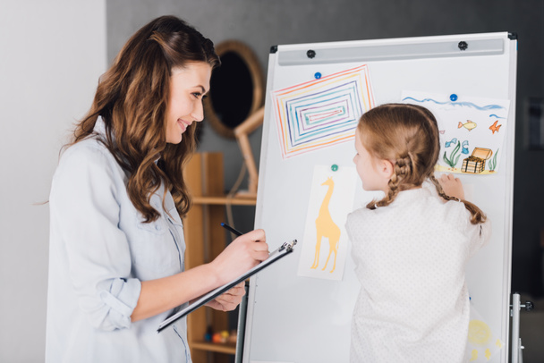 psicólogo feliz com prancheta conversando com a criança perto quadro branco com vários desenhos
 - Foto, Imagem