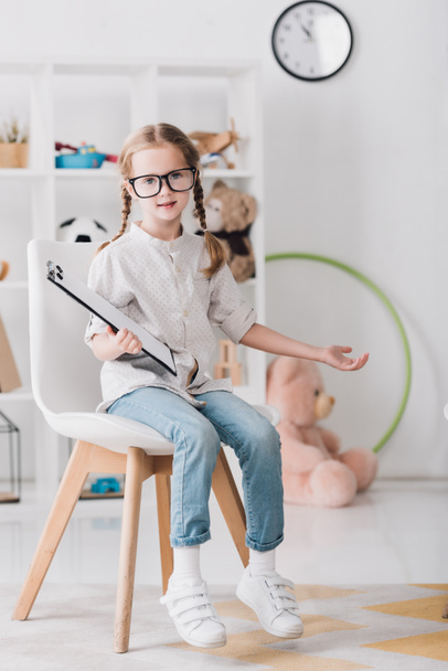 kindje in brillen met Klembord zittend op een stoel en camera kijken - Foto, afbeelding