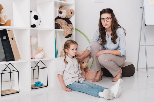 adult female psychologist supporting depressed child while she sitting on floor and looking at camera - Φωτογραφία, εικόνα