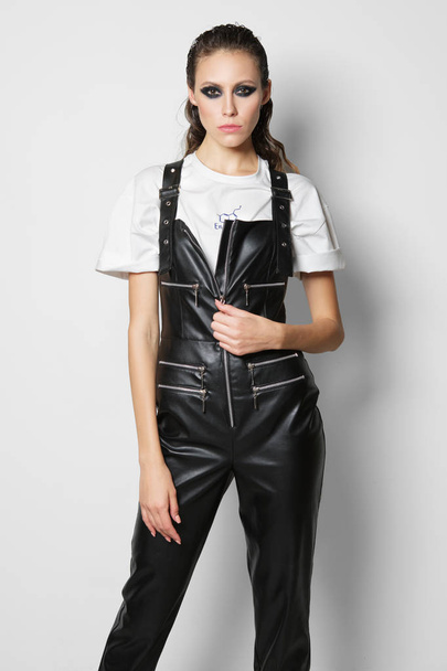 Girl in faux leather overalls and t-shirt posing near white wall - Foto, afbeelding