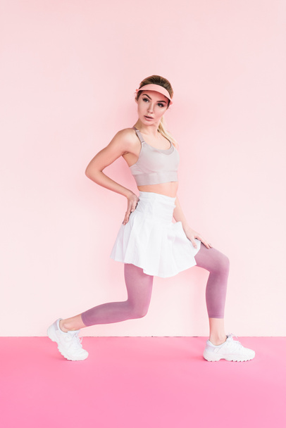 attractive stylish sportswoman in visor hat doing exercise on pink - Valokuva, kuva