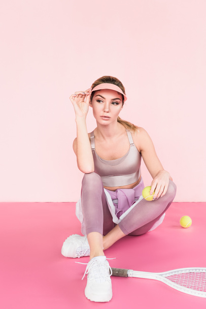 красивая спортсменка в козырьковой шляпе позирует с теннисной ракеткой и мячами на розовом
 - Фото, изображение