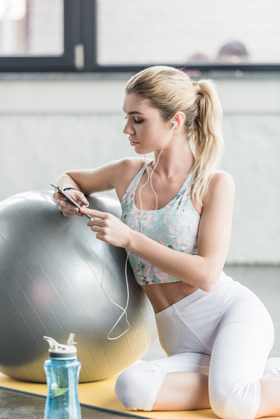 giovane bella donna in auricolare ascoltare musica con smartphone vicino palla fitness in palestra
 - Foto, immagini