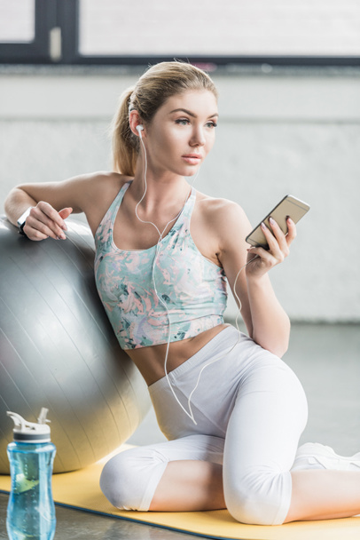 selective focus of female athlete in earphones resting and listening music with smartphone near fitness ball at gym - Photo, Image