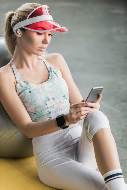 selective focus of sporty girl with smartwatch using smartphone near fitness ball at gym - Photo, Image
