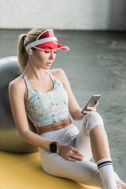 beautiful sportswoman with smartwatch using smartphone near fitness ball at gym - Photo, image