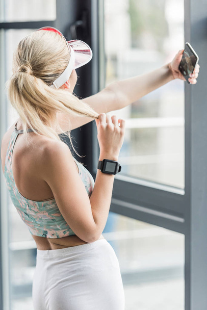 Widok z tyłu z sportsmenki z zegarka smartwatch biorąc selfie na smartfonie siłowni - Zdjęcie, obraz