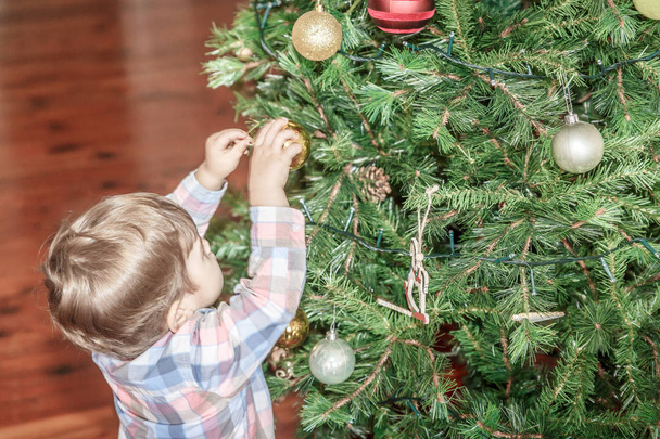 Sevimli küçük bir çocuk oturma odasında büyük bir Noel ağacına dekoratif bir top yerleştirir - Fotoğraf, Görsel