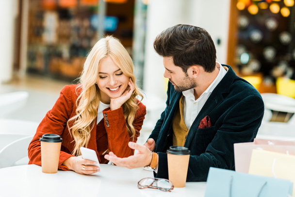 hermosa pareja joven utilizando el teléfono inteligente en la cafetería con café para ir y bolsas de compras
 - Foto, imagen