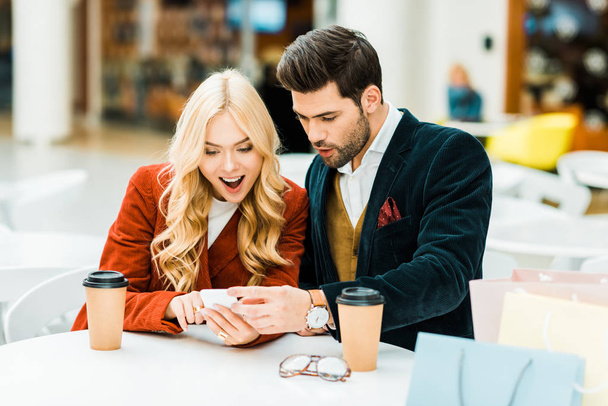 pareja emocionada utilizando el teléfono inteligente en la cafetería con café para ir y bolsas de compras
 - Foto, imagen