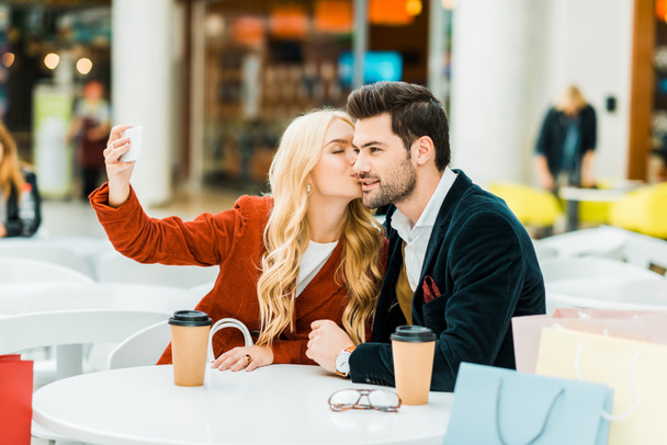 beautiful girl kissing boyfriend and taking selfie on smartphone in shopping mall  - Photo, Image