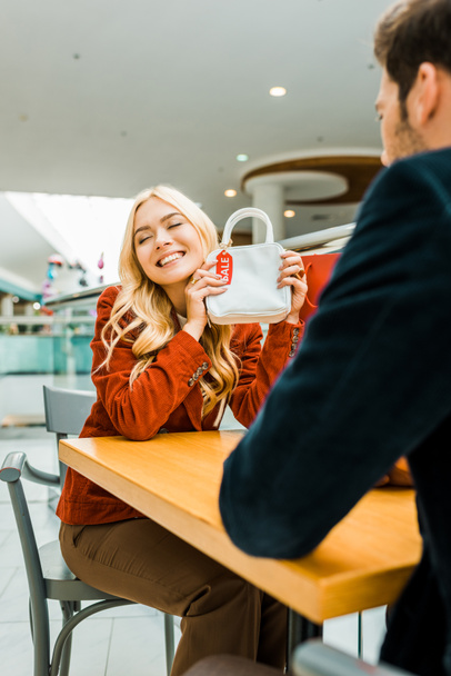 happy attractive woman showing bag with sale tag to boyfriend in shopping mall - Fotoğraf, Görsel