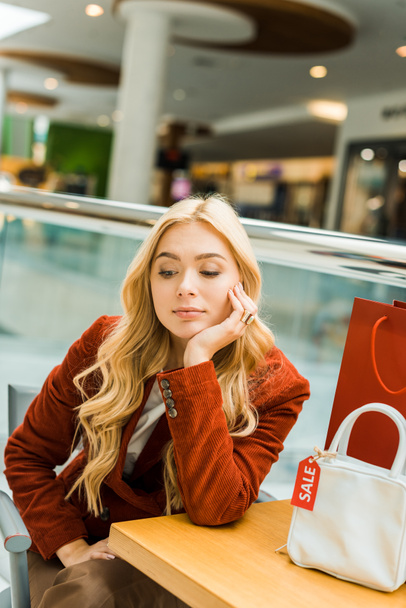 zdenerwowany blond dziewczyna siedzi w centrum handlowe z jednej torby na zakupy i jeden worek z tag sprzedaż   - Zdjęcie, obraz