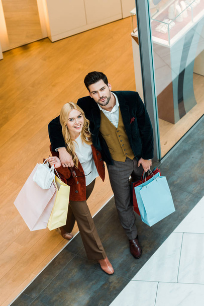 Blick von oben auf das schöne glückliche Paar, das sich umarmt und Einkaufstüten hält, während es aus der Boutique geht - Foto, Bild