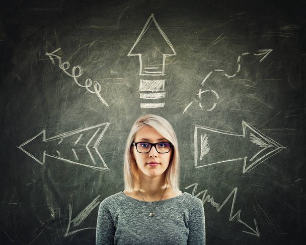 Young woman wearing glasses in front of a blackboard with arrows pointed to different directions. Difficult choice, decide which way to go, doubt concept. - Photo, Image