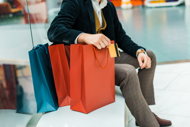 vista recortada del hombre con tarjeta de crédito y sentado con bolsas en el centro comercial
 - Foto, Imagen