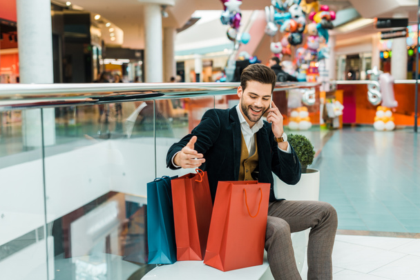 handsome fashionable man with shopping bags talking on smartphone while sitting in shopping mall  - Photo, Image