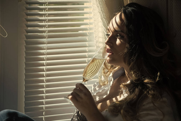 Девушка с длинными вьющимися волосами пьет шампанское у окна
 - Фото, изображение