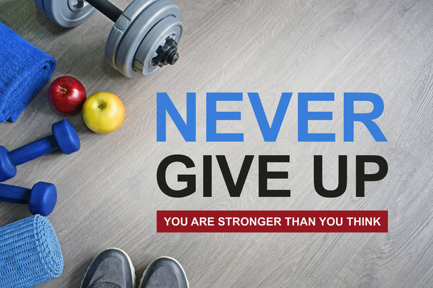 Never Give Up. Je bent sterker dan je denkt. Fitness motiverende citaten. Sport thema. Gezonde en actieve levensstijl concept. Sneakers, halters, appels en een training mat op grijze houten achtergrond.  - Foto, afbeelding