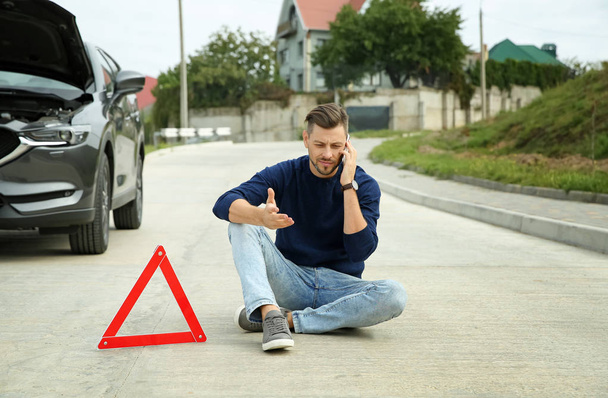 Homem falando no telefone perto de triângulo de aviso e carro quebrado na estrada
 - Foto, Imagem