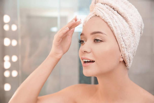 Beautiful woman with towel on head applying face cream in bathroom - Photo, Image