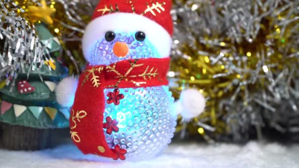 Bir kırmızı Noel şapka ve kırmızı eşarp üzerinde beyaz yün giyen parlayan bir dekoratif kardan adam rakam kar gibi yeşil Noel ağacı ve altın gelin teli ile arka plan üzerinde görünür. - Video, Çekim