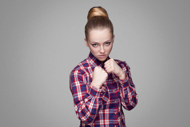 Serious beautiful blonde woman in pink checkered shirt with collected bun hairstyle looking at camera with boxing fists and ready to attack on gray background - Photo, Image