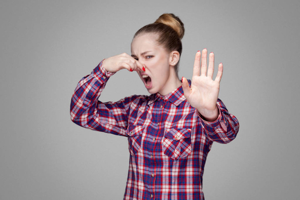 angry blonde woman in pink checkered shirt with collected bun hairstyle looking at camera with stop gesture and pinching her nose on gray background - Foto, Bild