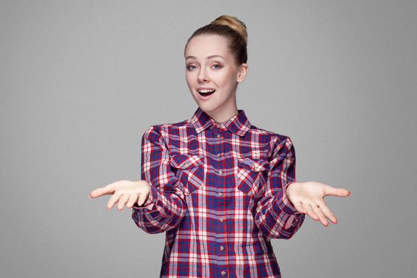 beautiful blonde woman in pink checkered shirt with collected bun hairstyle looking at camera with smile and raised arms and sharing on gray background - Φωτογραφία, εικόνα