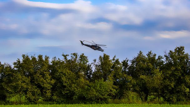 Helicóptero de combate en el cielo azul con nubes blancas. Árboles y arbustos con hierba
. - Foto, imagen