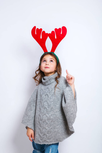 Happy little smiling girl showing product. Christmas concept. Smiling funny girl in deer horns in studio. - Photo, Image