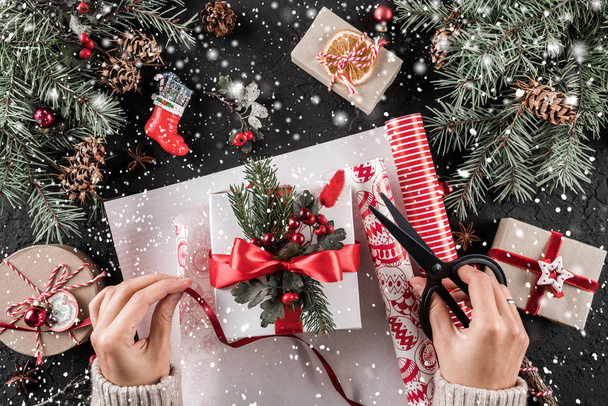 Die weiblichen Hände packen eine Weihnachtsgeschenkschachtel mit roter Schleife auf festlichem Hintergrund mit Tannenzweigen, Tannenzapfen. Weihnachten und ein frohes neues Jahr Karte, Bokeh, funkeln, glühen. flache Lage, Draufsicht - Foto, Bild