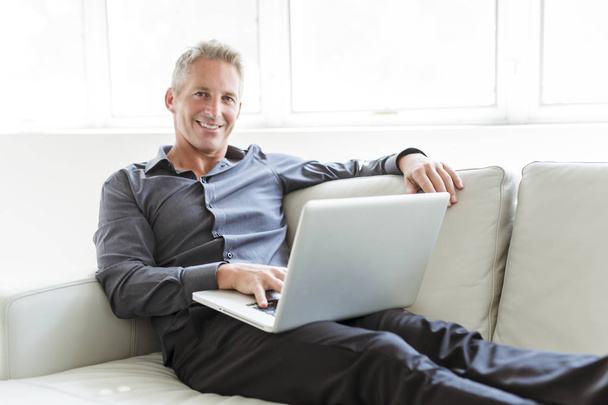 Portrait of happy mature man using laptop lying on sofa in house - Photo, image