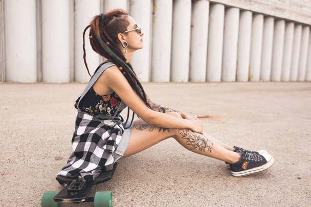 young girl with tattoo and dreadlocks sitting on a longboard on the background of concrete structure - Фото, изображение