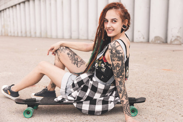 young girl with tattoo and dreadlocks sitting on a longboard on the background of concrete structure - 写真・画像