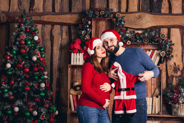 Σύζυγος και η σύζυγος του έγκυος κατέχουν ένα κοστούμι Χριστουγέννων για το μωρό. - Φωτογραφία, εικόνα