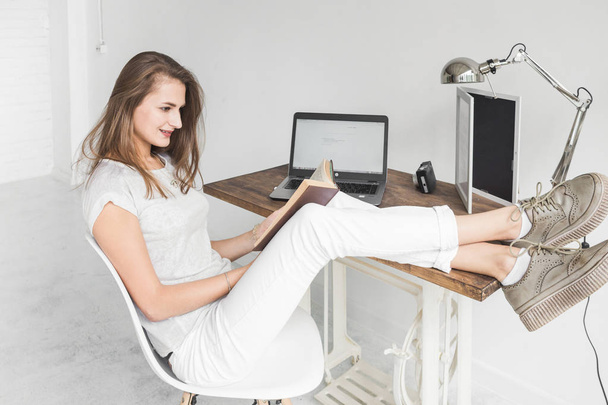 Young business woman working at home and reading a book with her legs on the table. Creative Scandinavian style workspace. - Photo, Image