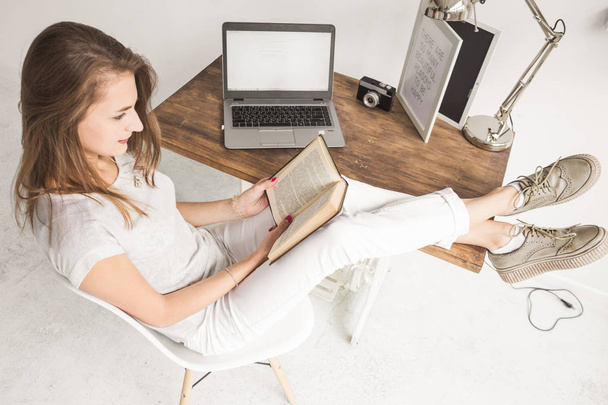 Young business woman working at home and reading a book with her legs on the table. Creative Scandinavian style workspace. Wiew from above - Photo, Image