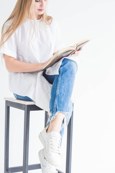 Portrait d'une jeune fille blonde élégante en T-shirt blanc et jean bleu lisant un livre sur fond blanc
 - Photo, image