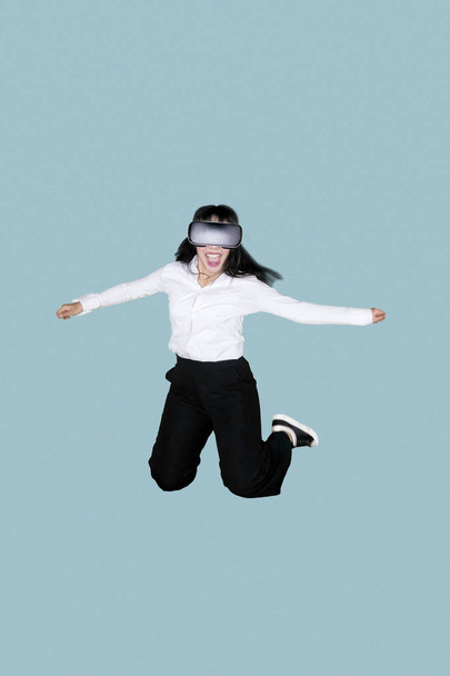 Portrait of a happy female entrepreneur wearing virtual reality goggles while jumping in the studio - Foto, imagen