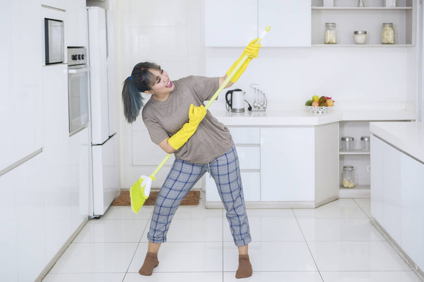 Portrait of happy housewife playing guitar with a broom while standing in the kitchen - Foto, imagen